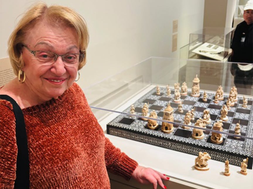 An older woman wearing glasses and a rust-colored sweater smiles while standing beside a glass-covered ornate chess set on display in a museum.