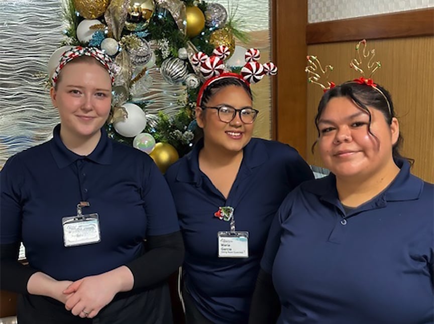 Three women wearing navy blue uniforms and festive headbands—one with peppermint candies and another with reindeer antlers—smile in front of a decorated Christmas tree with silver, gold, and white ornaments.