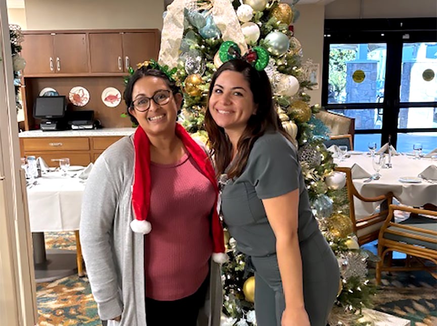 Two women smiling and posing in front of a decorated Christmas tree in a dining area. One wears glasses and a Santa hat scarf, and the other has festive green reindeer antlers. The room is set for a holiday meal.