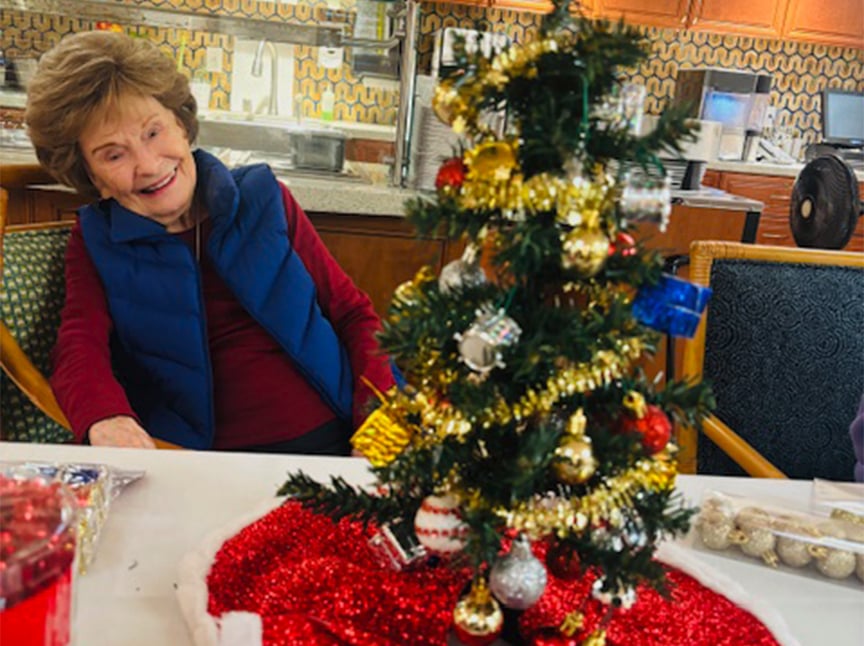 An elderly woman smiles while sitting at a table decorated with a small Christmas tree adorned with ornaments, tinsel, and gifts. The festive setting includes a red tablecloth and snacks.