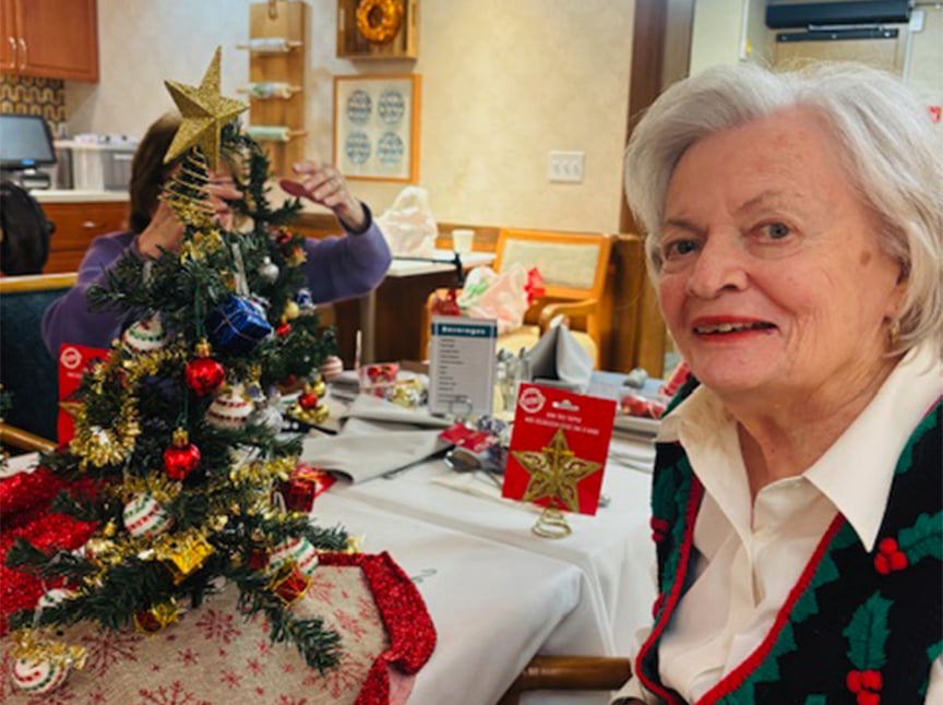 An elderly woman smiles next to a small decorated Christmas tree on a table. Another person adjusts the tree in the background. The setting appears festive and cozy, with holiday decor and wrapped gifts on the tables.