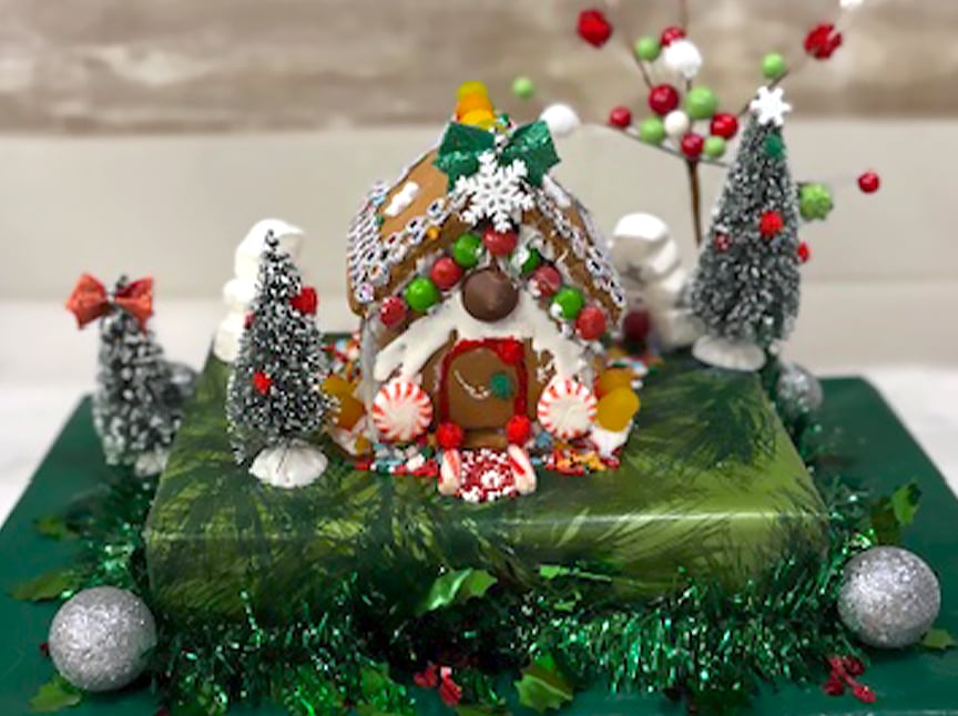 A decorated gingerbread house sits on a green platform, surrounded by snowy trees, ornaments, and colorful candy. The house has a candy-covered roof, peppermint decorations, and festive bows among wintry scenery.