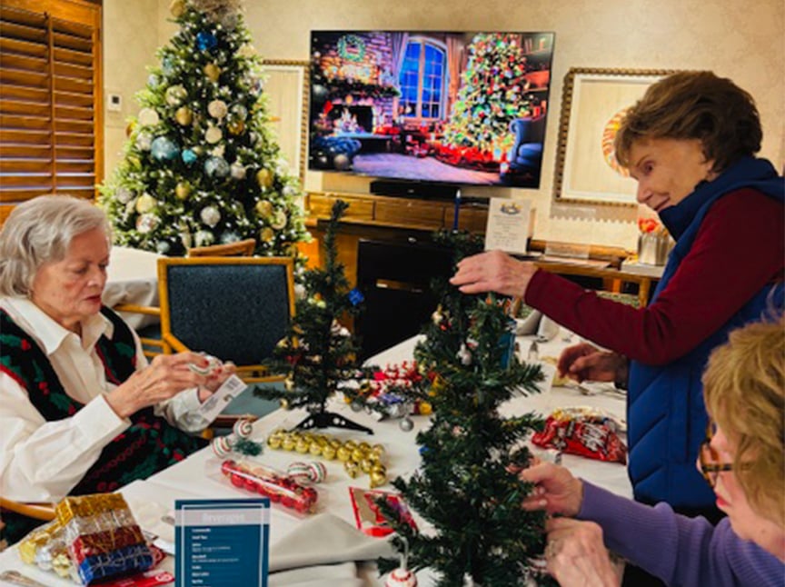 Three elderly women decorate miniature Christmas trees at a table, surrounded by holiday ornaments and gift boxes. In the background, a TV displays a festive fireplace scene and a decorated Christmas tree.