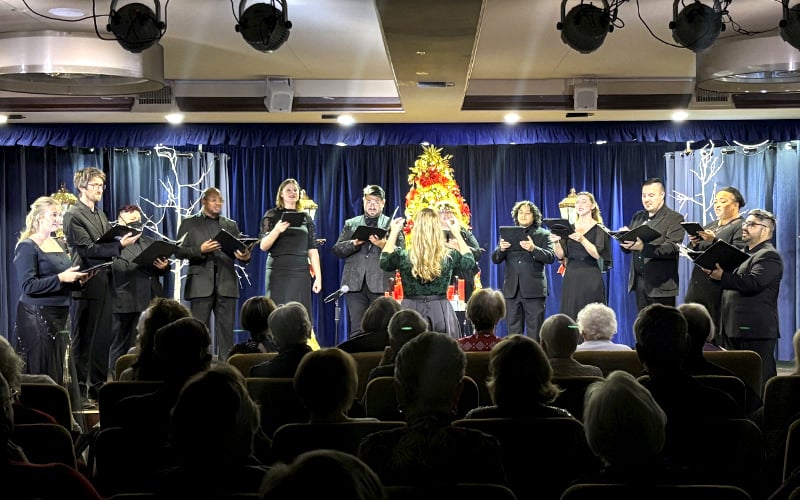 A choir dressed in black sings on stage directed by a conductor, with a decorated Christmas tree and blue curtains behind them. An audience is seated in front, watching the performance.