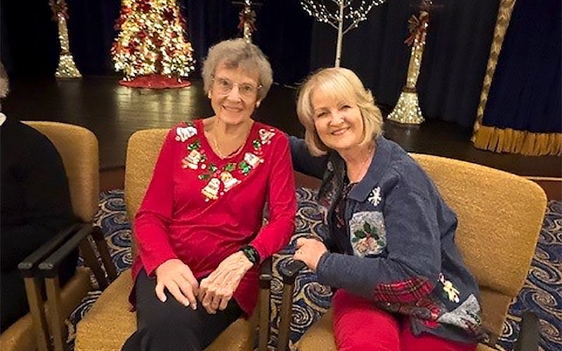 Two older women sit smiling together on tan chairs in a festive room with Christmas decorations, including a lit Christmas tree, in the background. They are wearing holiday-themed sweaters.