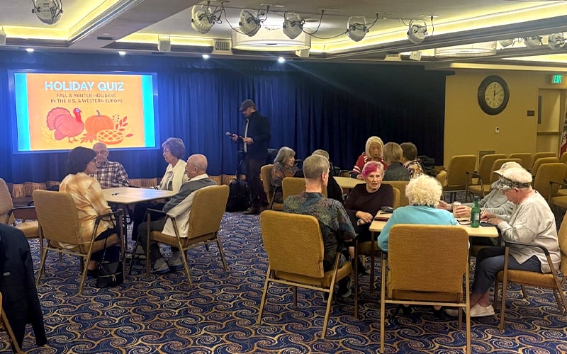A group of seniors sit at tables in a community room, participating in a holiday quiz event. A screen displays “Holiday Quiz” with festive graphics, while a man stands nearby with a microphone and notes.