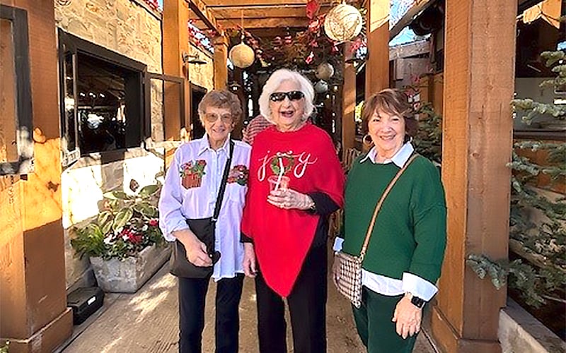 Three older women stand outside on a sunny day, smiling at the camera. They are dressed in festive holiday clothing, with Christmas decorations and greenery in the background.