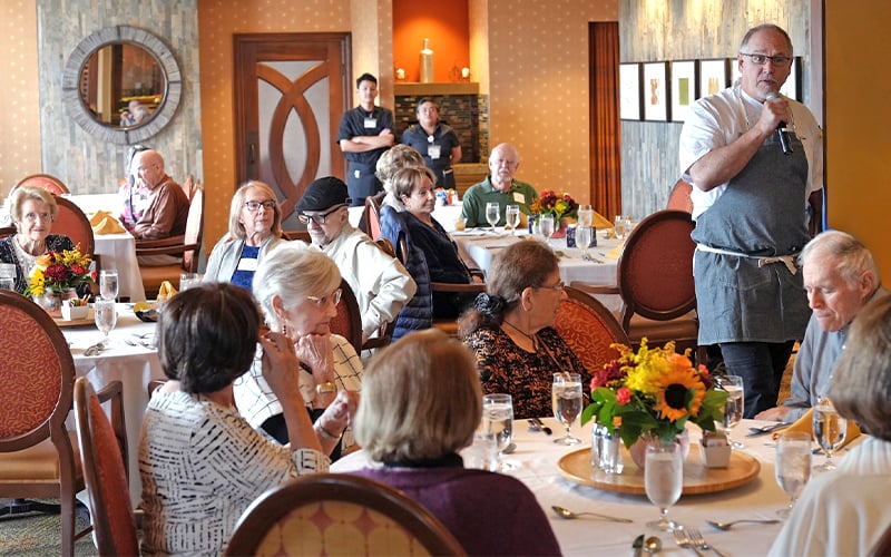 A man stands and speaks with a microphone to a group of older adults seated at round tables in a restaurant or dining hall, decorated with flowers and warm lighting. Two staff members stand in the background.
