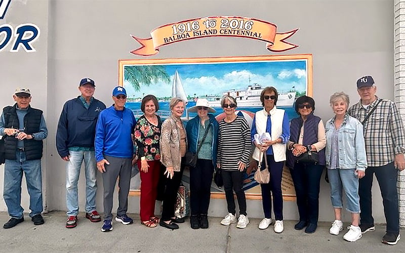Ten adults stand in a row, smiling in front of a mural celebrating the Balboa Island Centennial (1916–2016), which features a boat and waterfront scene. Most are wearing casual clothing, sunglasses, and jackets.