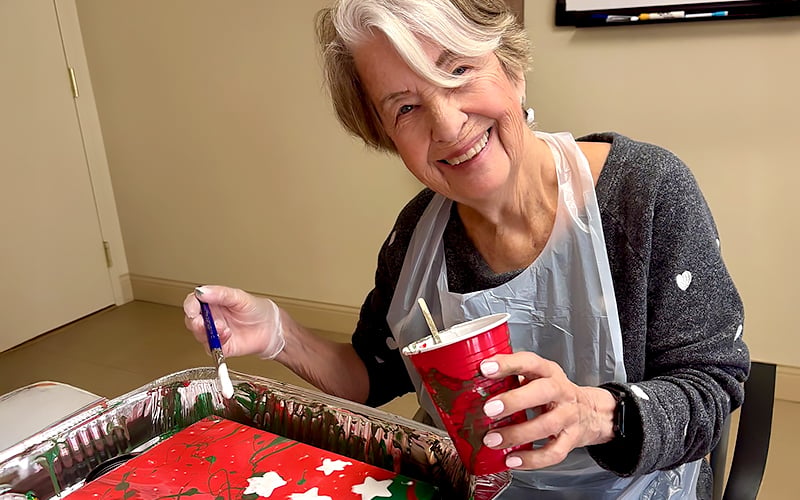 An older woman with gray hair smiles while decorating a red holiday-themed tray. She wears a gray sweater with white hearts, a plastic apron, and holds a paintbrush and a red cup, sitting at a table indoors.