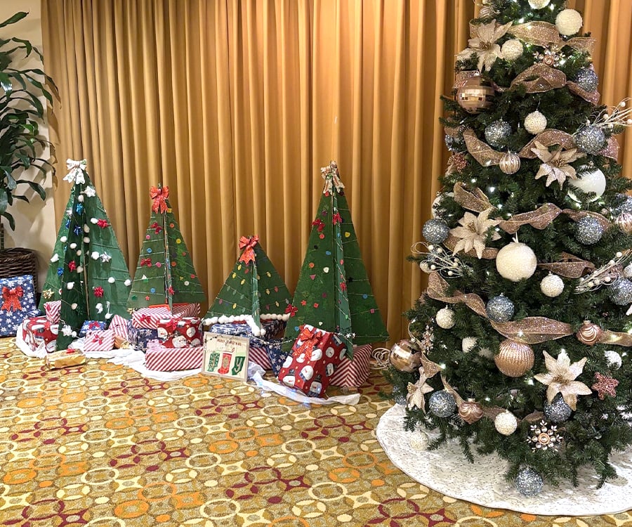 A decorated Christmas tree with ornaments and ribbons stands next to several green, triangular, cardboard trees with bows and stars. Wrapped presents are placed around the trees on a patterned carpet.