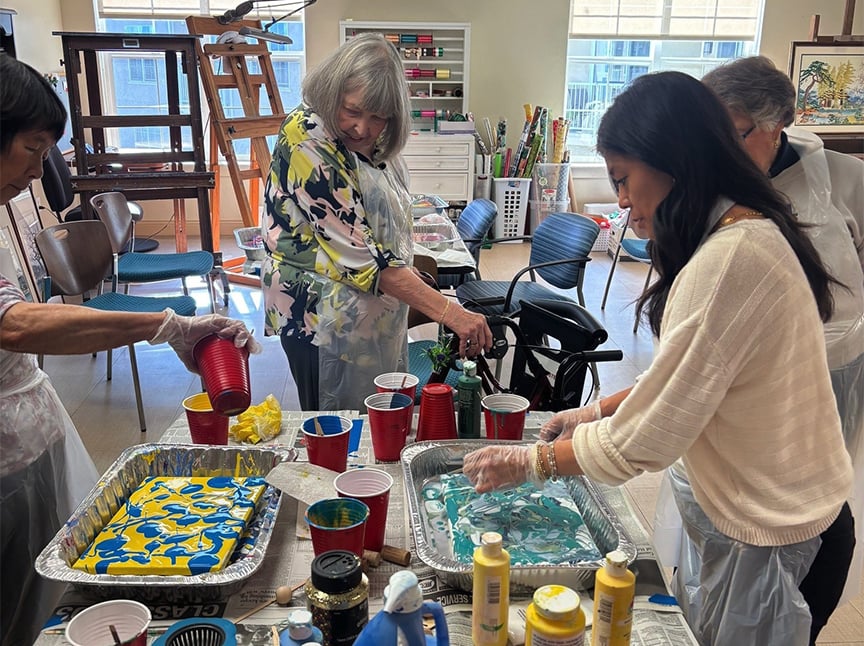 A group of women wearing gloves and aprons gather around tables with trays of paint, pouring colorful paint for an art project in a bright, well-lit art studio.