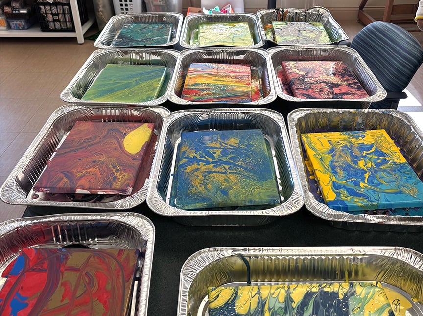 Several foil trays are arranged on a table, each holding a flat piece of paper or canvas with colorful, swirling paint patterns, likely created using a paint-pouring or marbling technique.