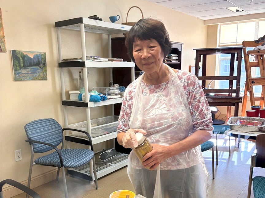 An older woman wearing a floral shirt and a plastic apron stands in a bright room, holding a spice jar. Shelves, chairs, and art supplies are visible in the background.