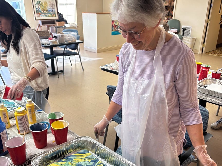 An older woman in a striped shirt and apron smiles while working on a marbling art project, surrounded by colorful paints and cups on a table in a bright, spacious room. Another person is painting in the background.
