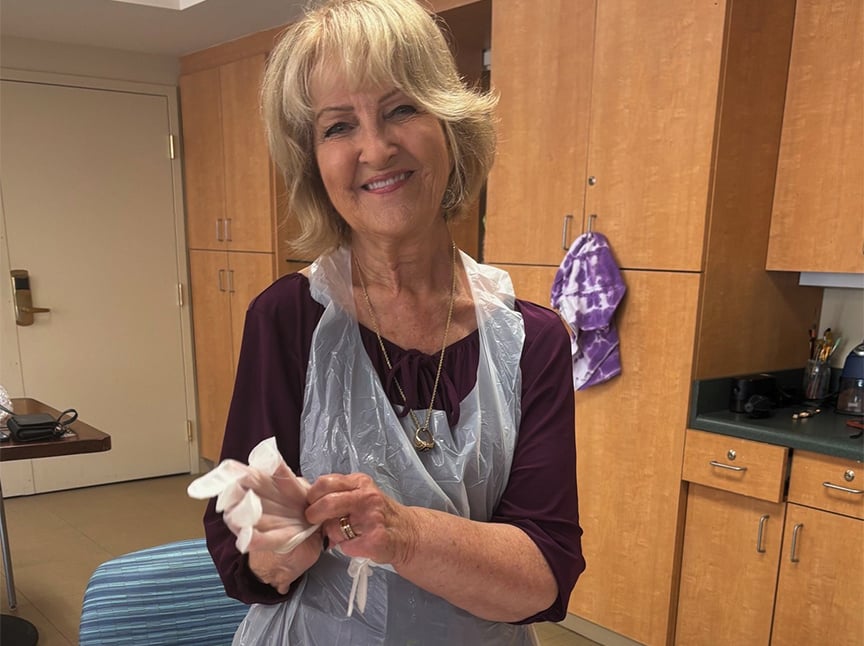 A smiling older woman with blond hair wears a plastic apron and is putting on disposable gloves in a kitchen with wooden cabinets.