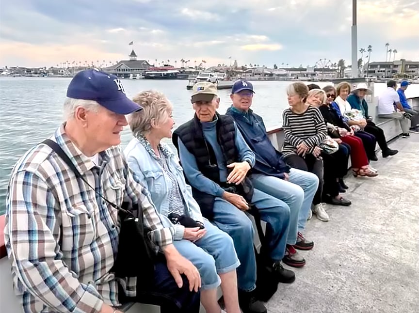 A group of older adults sit on a long bench by the waterfront, smiling and conversing. Boats and buildings are visible across the water under a cloudy sky.