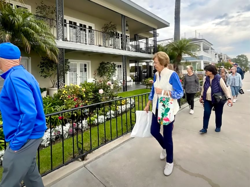 A group of people walk along a sidewalk next to a well-kept garden and a two-story building with balconies and flowers, under a partly cloudy sky.