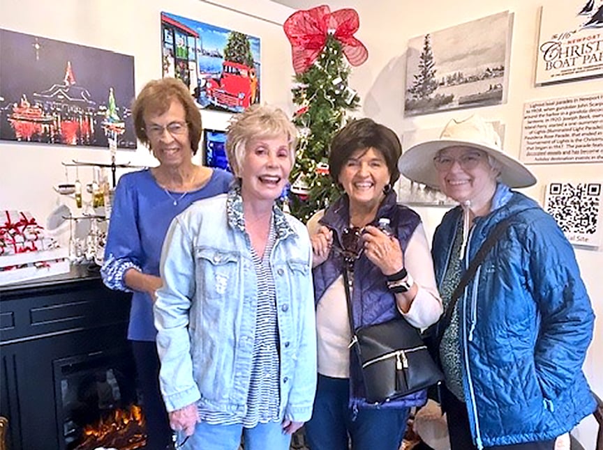 Four smiling women stand together indoors near a decorated Christmas tree, with art and informational signs on the wall behind them. They are dressed in casual clothes and appear to be enjoying a festive event.