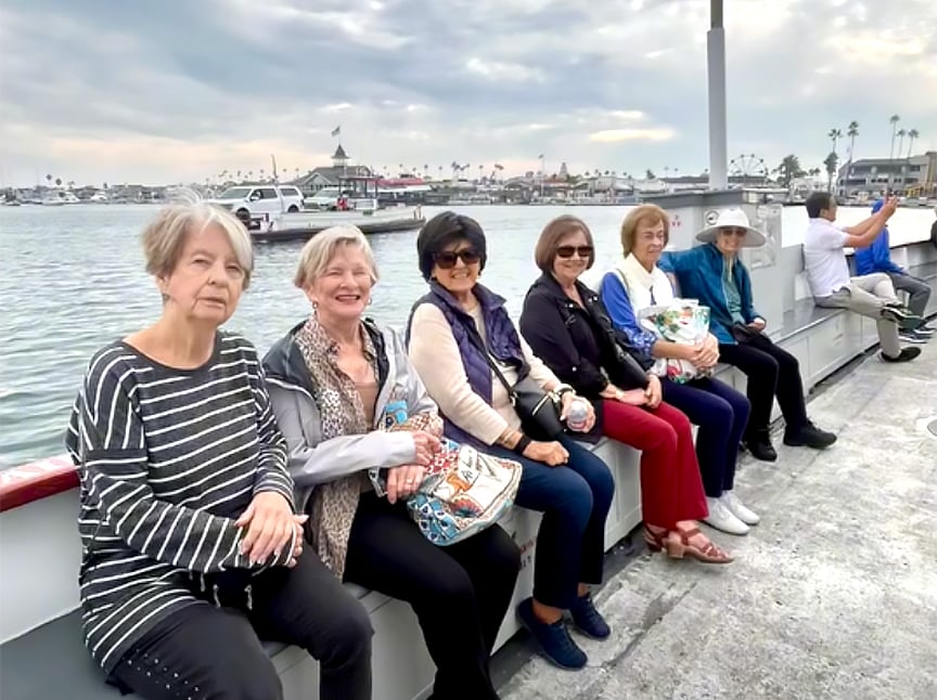 Six older women sit on a bench by the water, smiling at the camera, with a cloudy sky, boats, and a marina in the background. Another person sits at the far end, gazing away from the group.