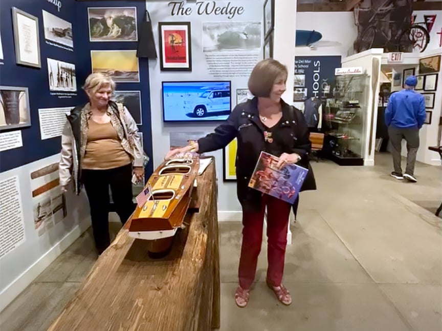 Two women stand in a museum exhibit; one touches a wooden boat model on display, while the other stands beside her. Museum walls feature photos, text, and artifacts, with more exhibits visible in the background.