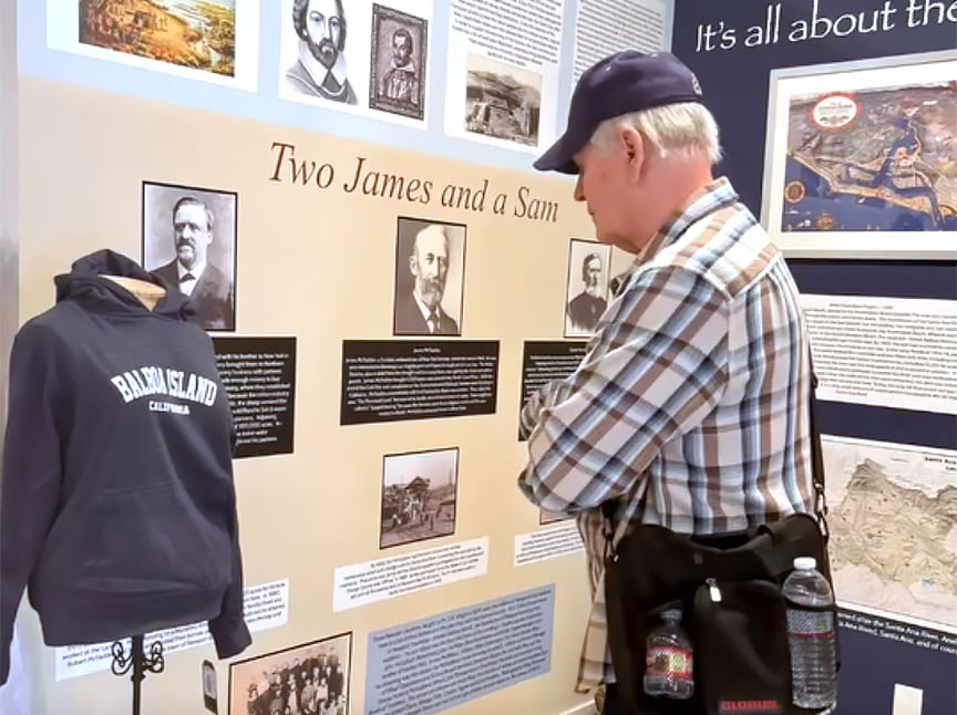 An older man in a plaid shirt and cap reads a museum exhibit titled Two James and a Sam, which displays historical photos, text, and a mannequin wearing a Balboa Island California hoodie.