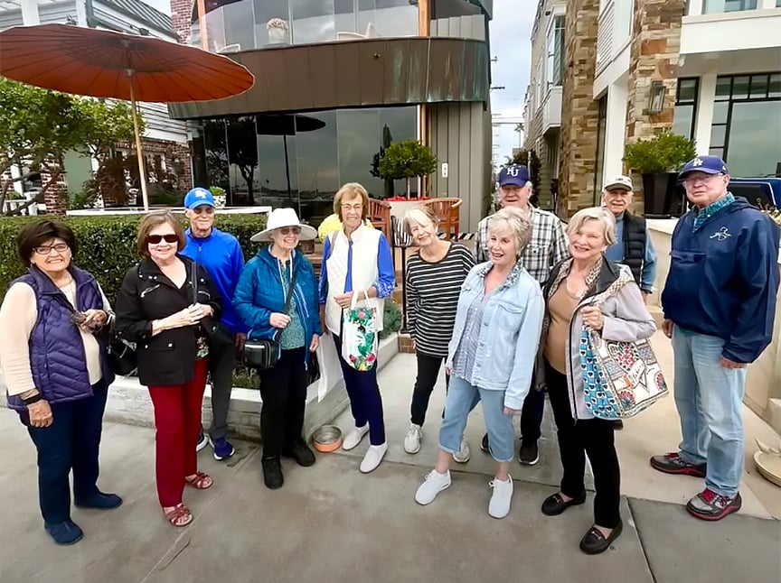 A group of twelve smiling adults, casually dressed, stand together outdoors on a sidewalk by a modern building. Some hold bags, and a couple wear hats. The atmosphere appears friendly and relaxed.