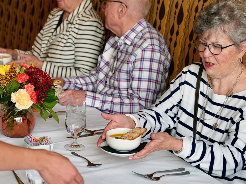 An older woman in a striped shirt receives a bowl of soup with bread at a restaurant table, sitting with two others. A vase of flowers, water glasses, and cutlery are on the white tablecloth.