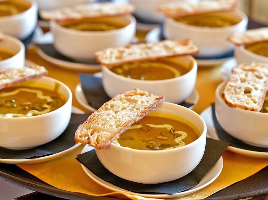 Several white bowls filled with soup are arranged on black napkins, each topped with a slice of crusty bread. The soup appears creamy with a drizzle of oil and seeds for garnish.