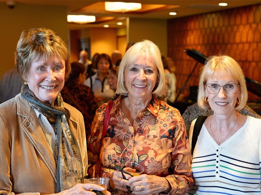 Three older women smiling and standing close together at an indoor social gathering. One holds a drink. The background shows other people, warm lighting, and textured walls, creating a friendly, welcoming atmosphere.