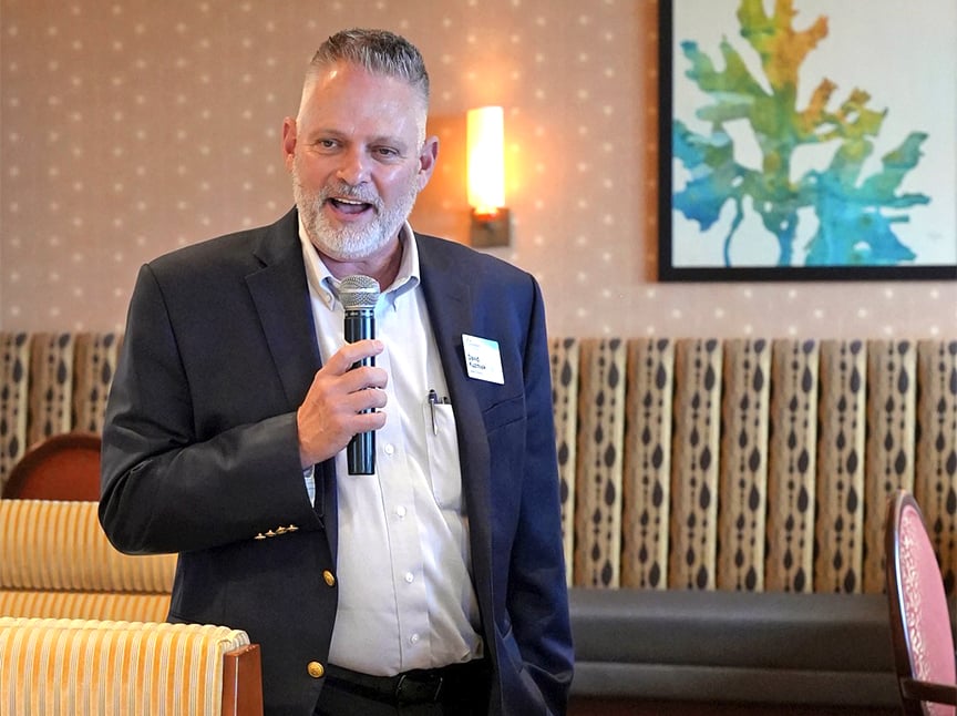 A man in a suit jacket holds a microphone and speaks in a room with patterned wallpaper and a colorful abstract painting on the wall behind him.