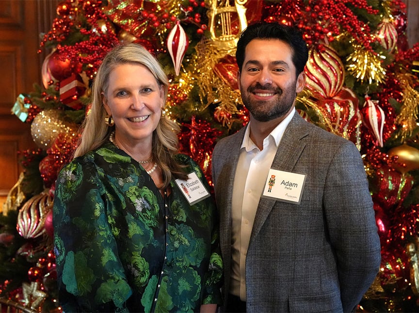 A woman and a man, both wearing name tags, stand smiling in front of a decorated Christmas tree with red and gold ornaments.