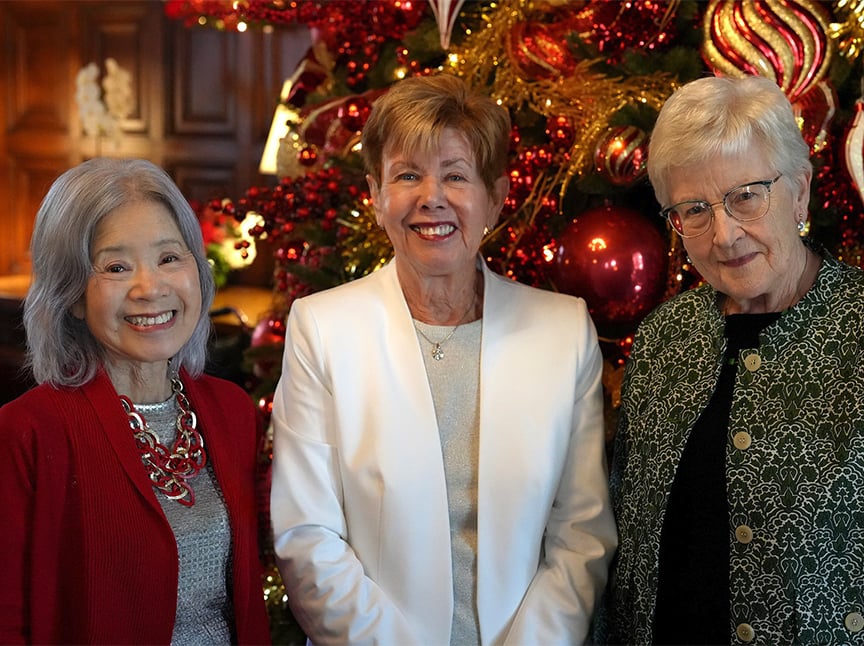 Three older women stand smiling in front of a decorated Christmas tree with red and gold ornaments. One wears a red dress, another a white blazer, and the third a green patterned jacket and glasses.