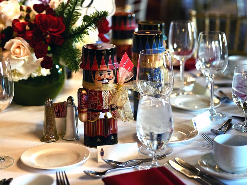 A festive table setting features glassware, plates, and silverware, with a nutcracker figurine and a gold ribbon as a centerpiece. A bouquet of red and white flowers decorates the table in the background.