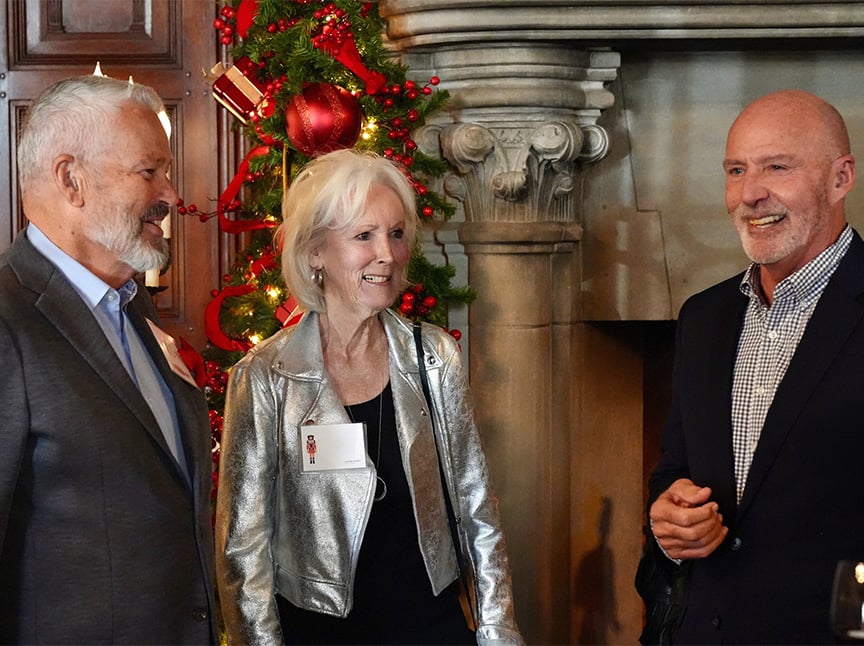 Three older adults, two men and one woman, are smiling and talking together indoors. Behind them is a decorated Christmas tree and an ornate fireplace, giving a festive atmosphere.