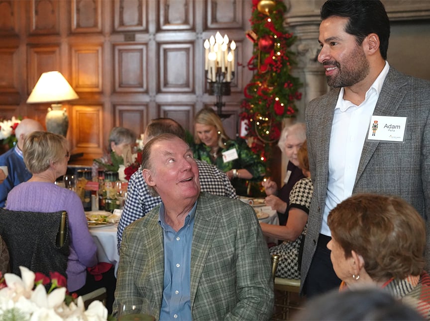 A man stands and smiles while talking to a seated man at a festive gathering. Other people are seated at tables, and a decorated Christmas tree is visible in the background.