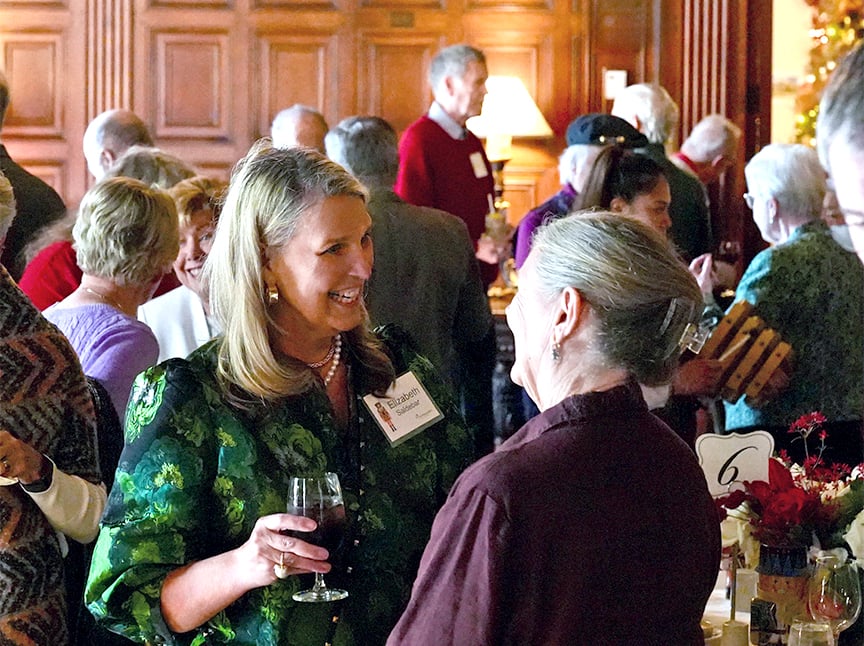 A group of people socializing at an indoor event. Two women in the foreground are smiling and talking; one holds a glass of red wine. Others mingle in the background in a warmly lit, wood-paneled room.