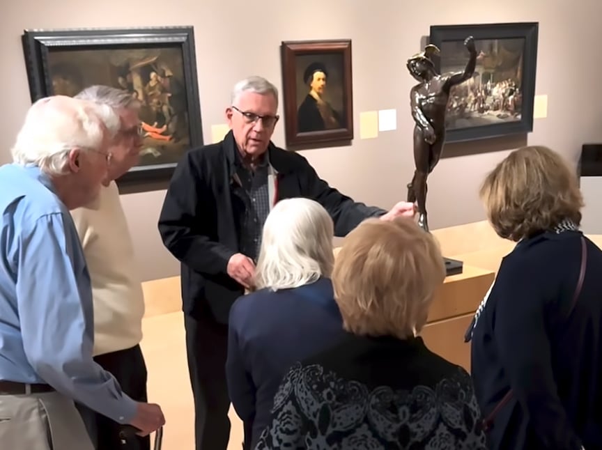 A museum guide explains a bronze statue to a group of older adults gathered around, with framed paintings displayed on the beige walls behind them.