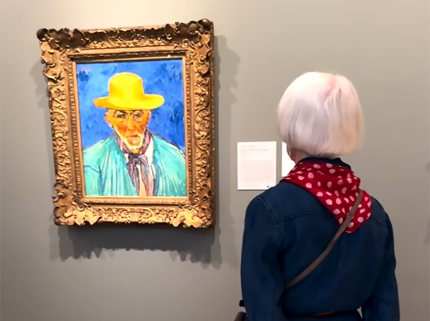 An older woman with white hair and a red polka-dot scarf views a colorful portrait of a man in a yellow hat, framed in ornate gold, hanging on a museum wall.