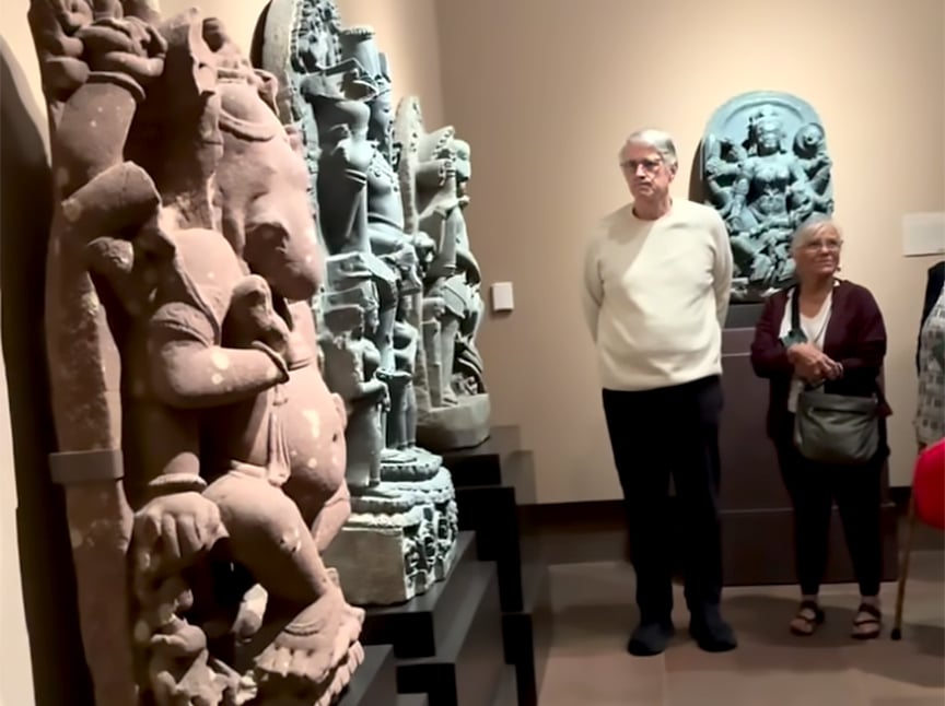 Two elderly people stand in a museum, observing large stone sculptures of Hindu deities displayed on pedestals. The background features another statue mounted on the wall.
