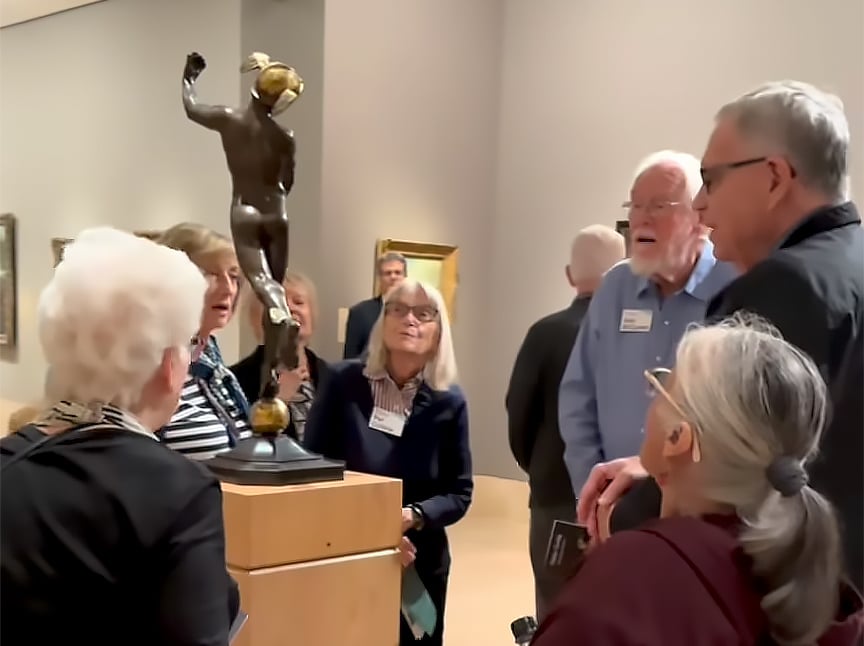 A group of older adults gathers around a bronze statue in an art gallery, engaging in conversation and observing the artwork. Several people wear name tags, and framed paintings hang on the walls in the background.