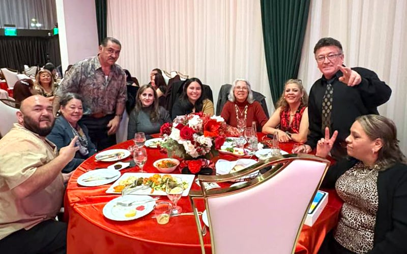 A group of people sit and stand around a round table covered with a red tablecloth, enjoying a meal. They are smiling and posing for the photo in a festive setting with floral centerpieces and draped curtains.