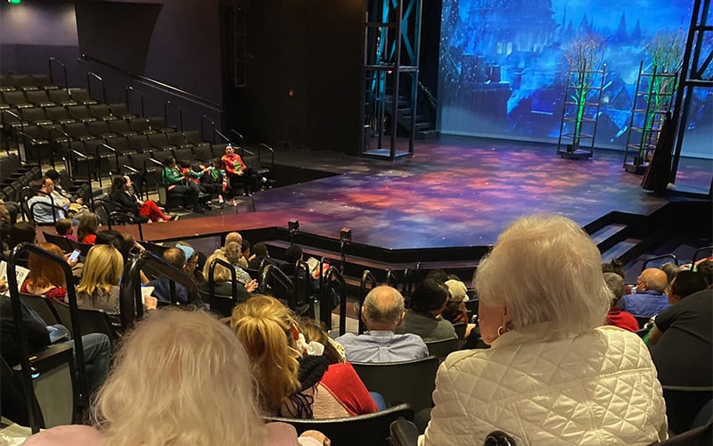 Audience members sit in a theater facing a stage with colorful lighting and a scenic backdrop, waiting for a performance to begin. The seats are partially filled and the atmosphere is anticipatory.