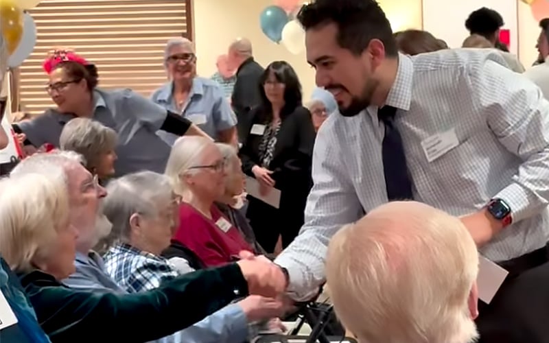 A man in a dress shirt and tie shakes hands with an elderly woman seated among a group of seniors at a lively indoor event. Other attendees and staff are smiling and interacting in the background.