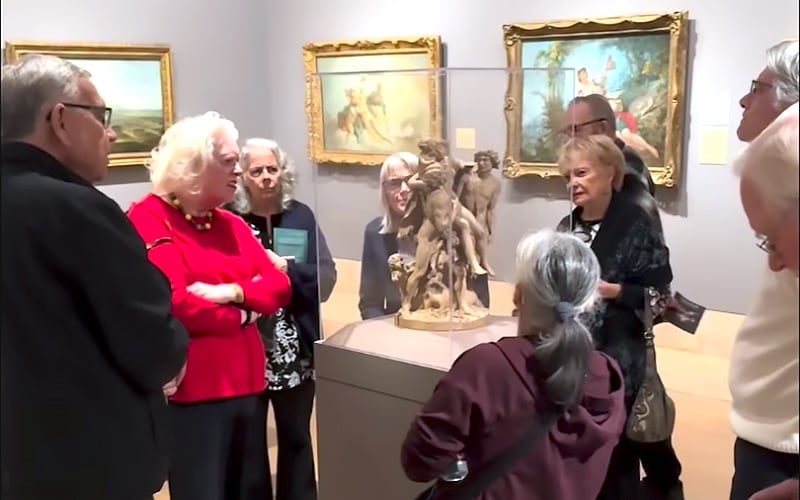 A group of older adults gathers around a sculpture in an art museum, listening intently to a woman in a red sweater. Framed paintings hang on the light gray walls in the background.