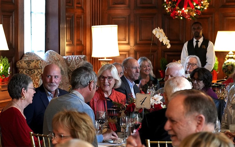 A group of older adults sit at round tables, smiling and conversing at an elegant event with wood-paneled walls and holiday decorations; a server stands in the background.