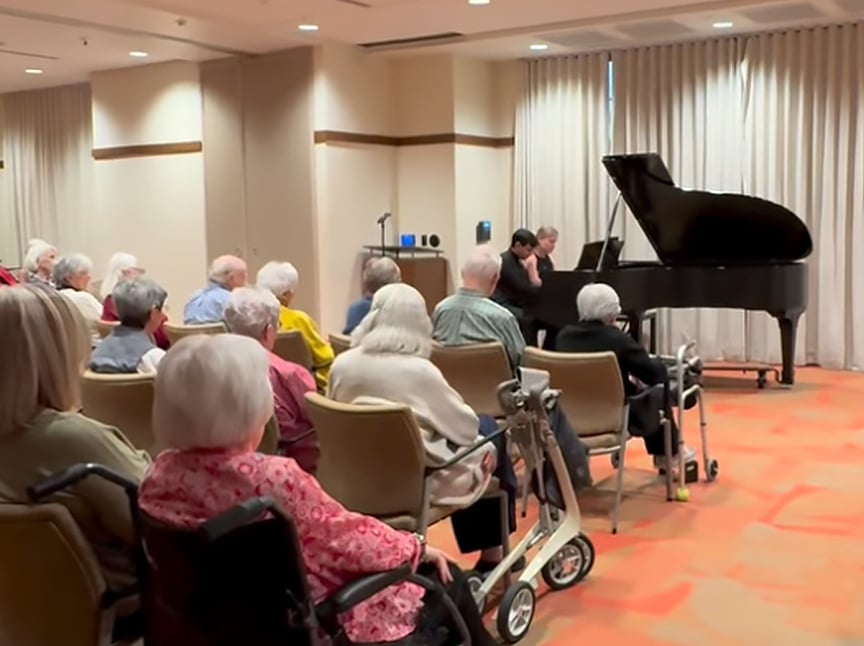 A group of elderly people sit in chairs and wheelchairs, watching a pianist perform on a grand piano in a brightly lit room with beige walls and curtains.