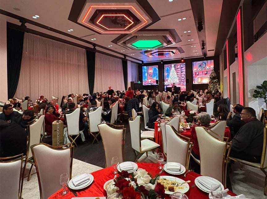 A large banquet hall filled with people seated at round tables with red tablecloths, festive decorations, and floral centerpieces. Large screens display holiday scenes, and the room is brightly lit with elegant chandeliers.