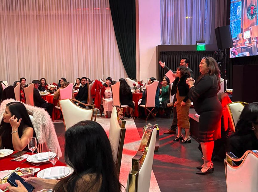 A group of people in formal attire sit and stand around decorated tables at a festive indoor event. Some are watching and reacting to something out of frame, while others are engaged in conversation. Red and white decor is visible.