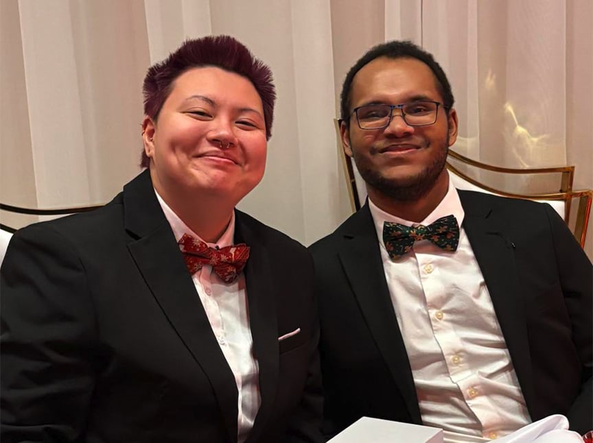 Two people dressed in black suits, white shirts, and patterned bow ties are smiling while seated indoors against a white curtain background.
